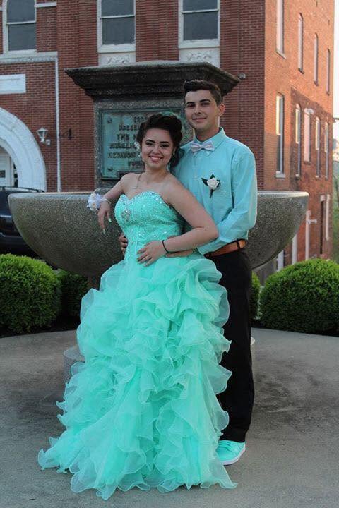 A couple poses outside a brick building, with the woman in a turquoise ruffled gown and the man in a matching shirt and bow tie.