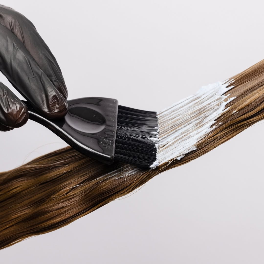 A gloved hand uses a black brush to apply white product to a section of brown hair.