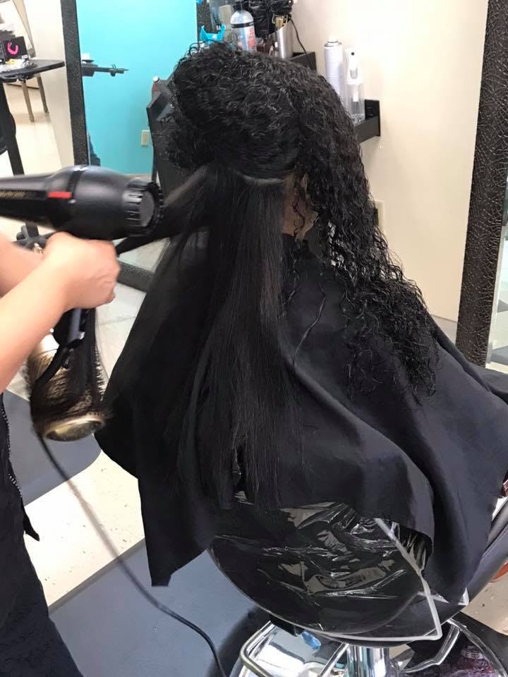 A hairstylist blow-dries a client’s sectioned, curly black hair in a salon, with the straightened lower section hanging beneath the curls.
