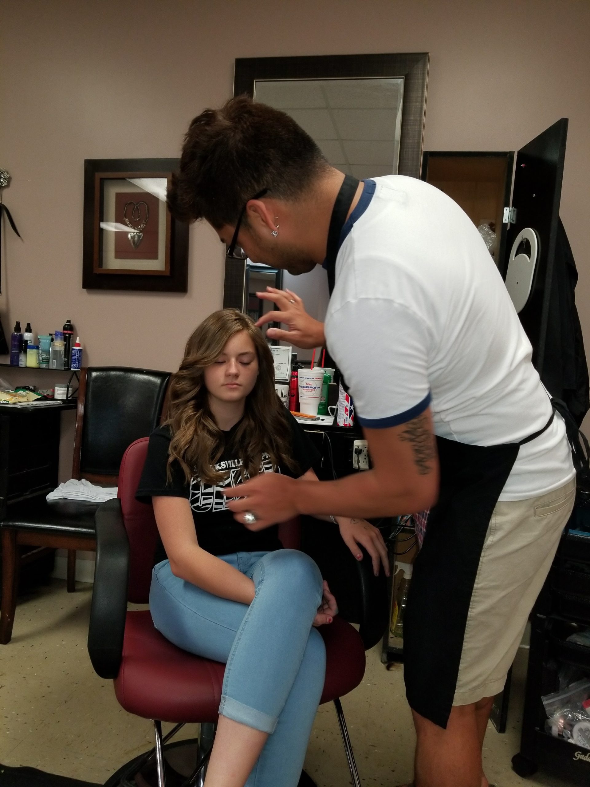 A man in a white shirt leans over a seated young woman in a salon chair, gesturing near her hair.