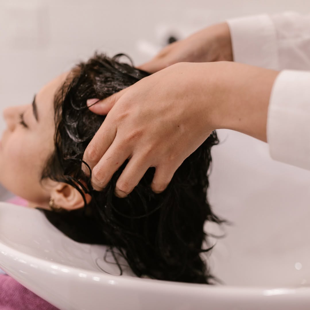 A person reclines at a salon sink while a stylist massages and washes their long dark hair.