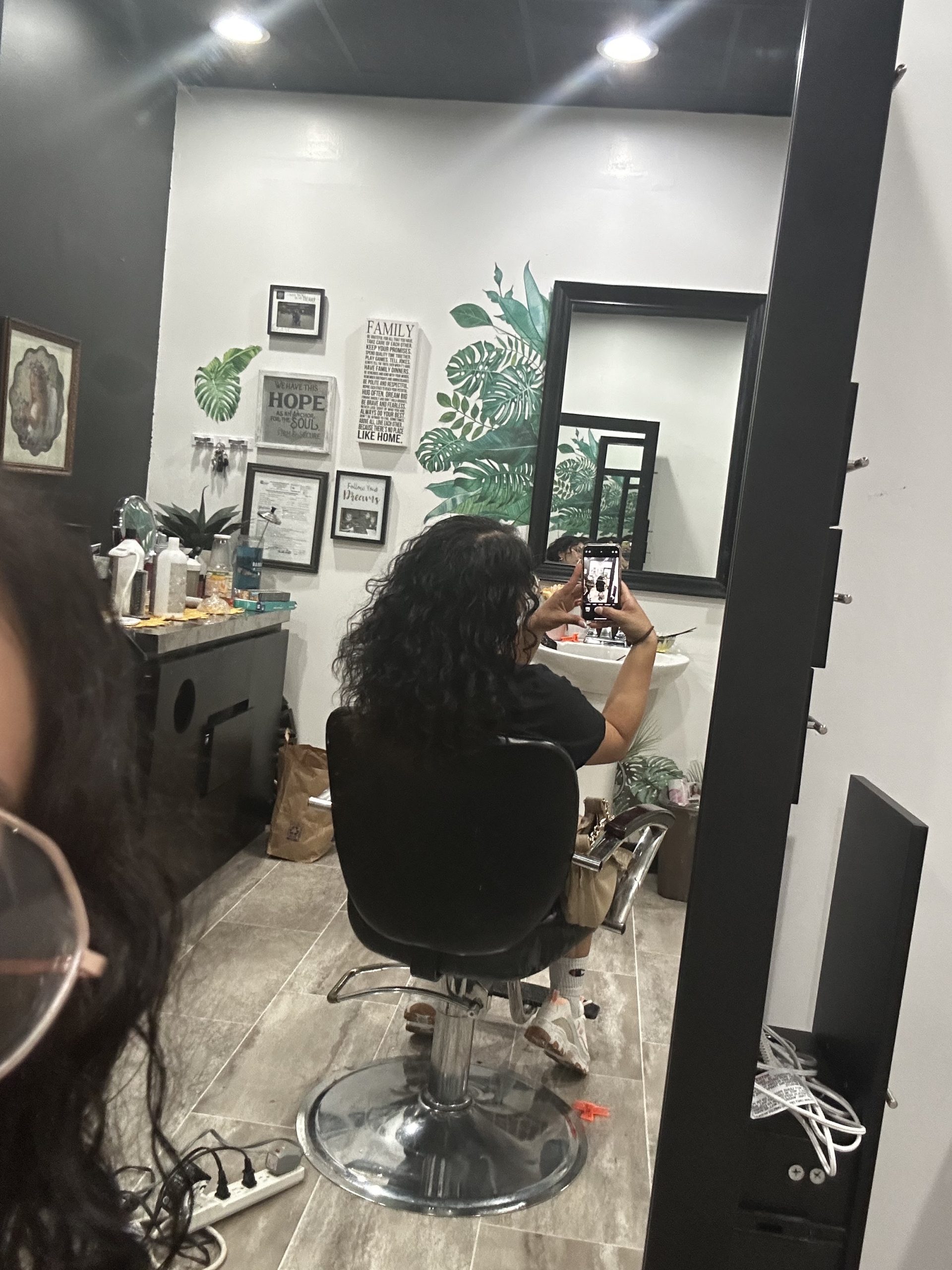 A person sits in a salon chair taking a phone selfie, with a wall mirror, hair products, framed art, and leafy plant murals behind them.