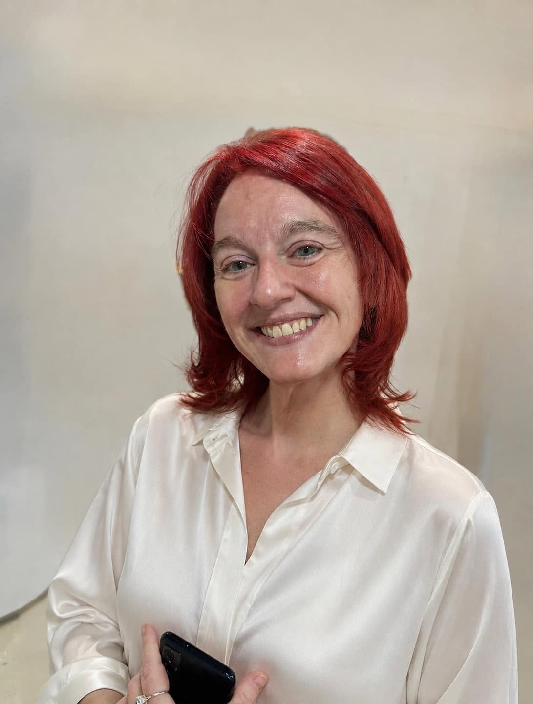A smiling red-haired woman in a white blouse holds a black phone against a plain light-colored wall.