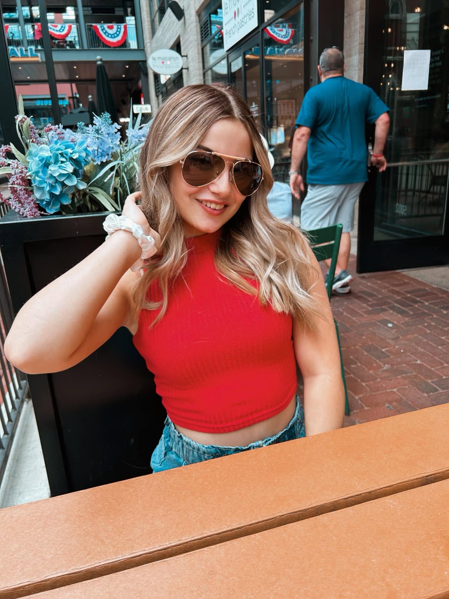 A smiling woman in sunglasses and a red sleeveless top sits at an outdoor restaurant table beside blue flowers.