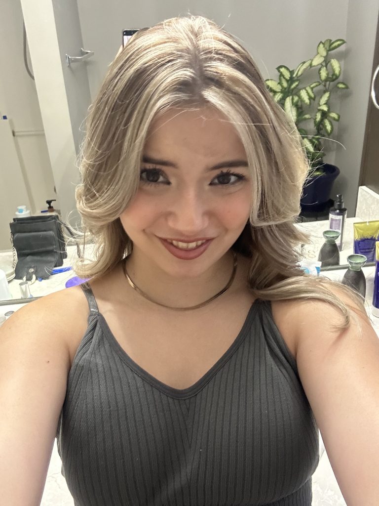 A smiling woman with shoulder-length blonde hair takes a selfie in a bathroom, wearing a dark ribbed tank top with toiletries and a plant behind her.