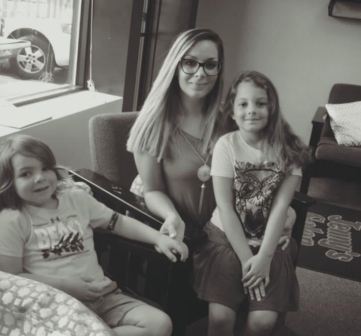 A woman wearing glasses sits between two young girls in a living room, with a car visible through the window.
