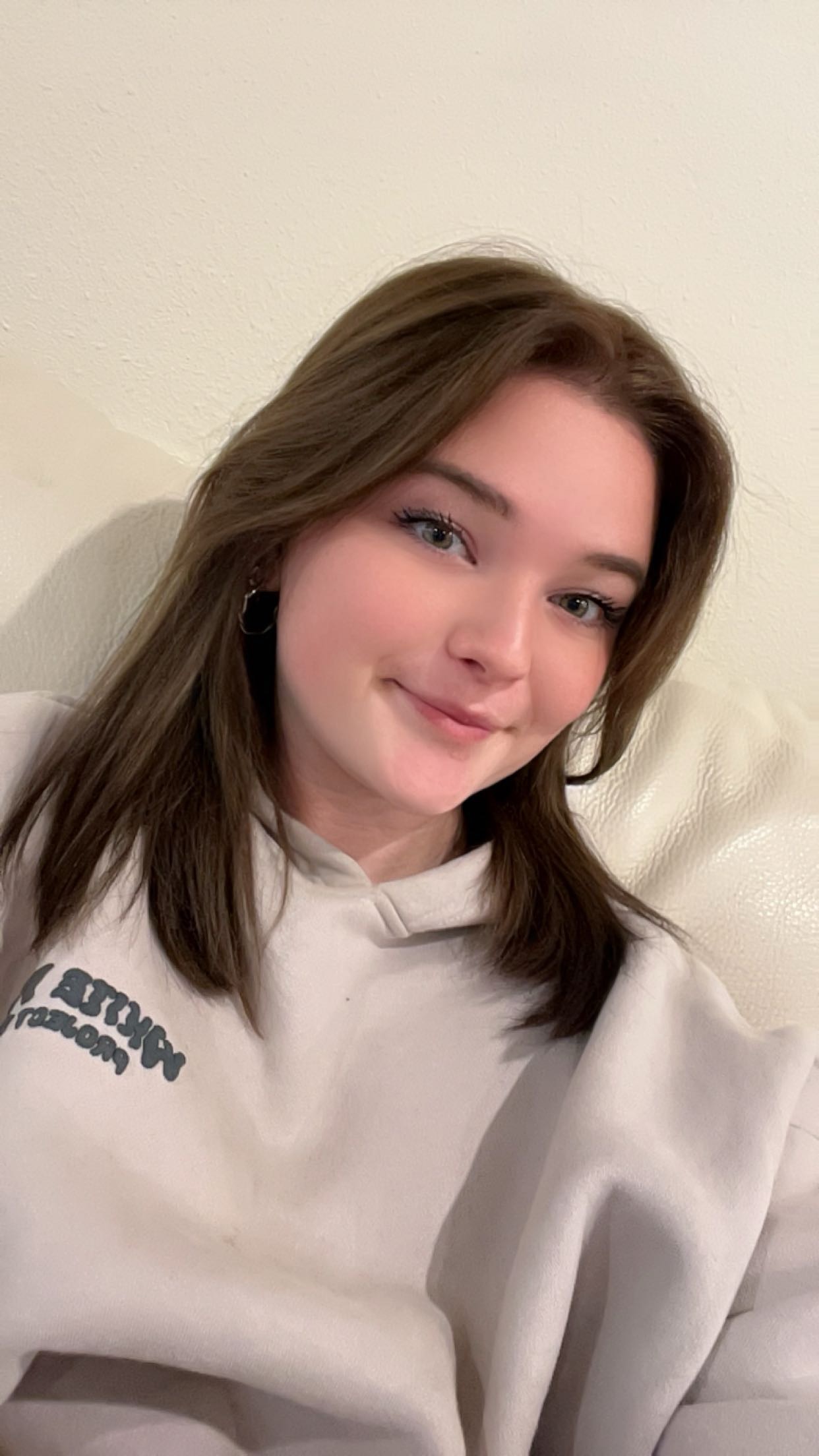A young woman with long brown hair smiles at the camera while sitting against a light-colored couch, wearing a white sweatshirt.