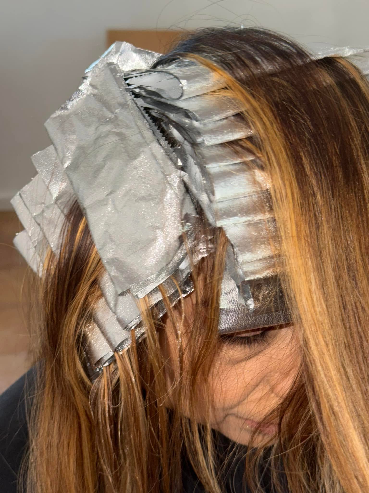 Close-up of a person with long brown hair sectioned and covered in numerous silver foil strips during a hair-coloring treatment.