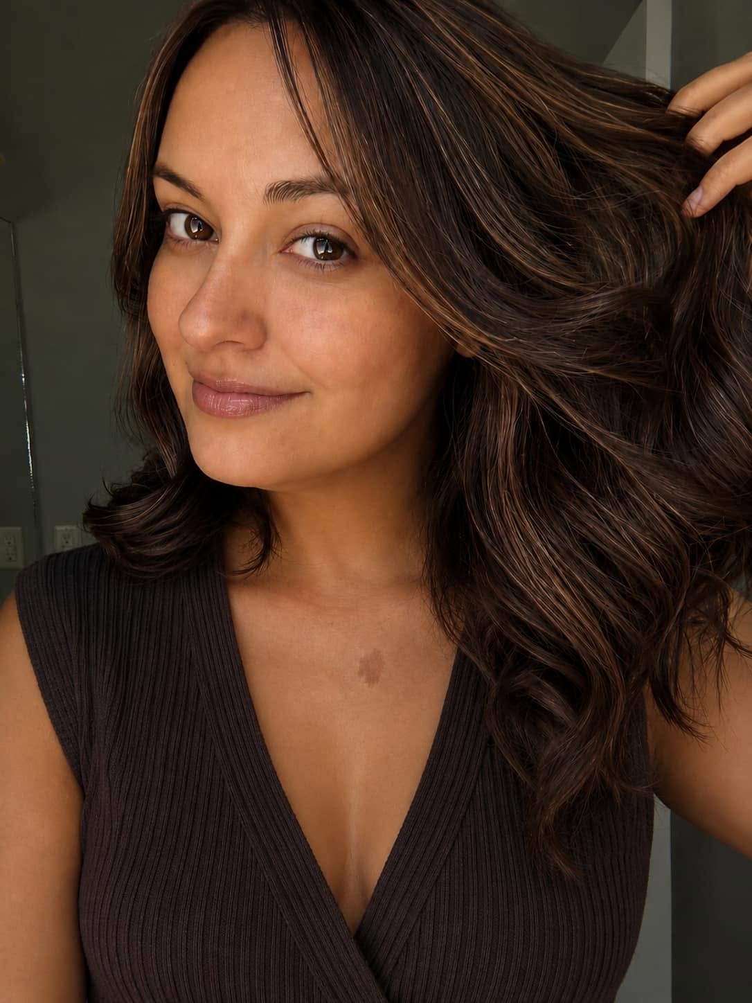 Close-up of a woman with wavy brown hair, wearing a dark ribbed sleeveless top, gently holding her hair and smiling softly.