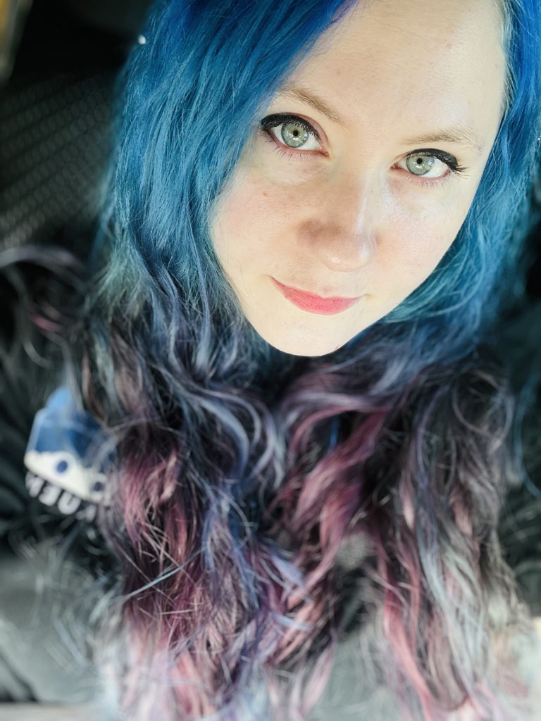 Close-up portrait of a person with bright blue hair, green eyes, red lipstick, and long wavy hair streaked with purple and pink.